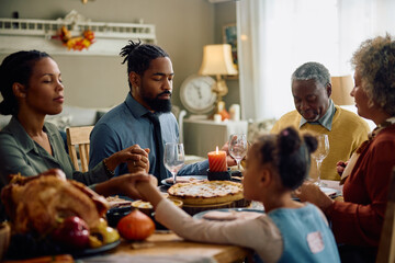 Black man and his extended family praying during Thanksgiving meal at dining table.
