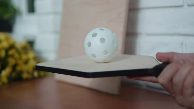 Player tests craft wooden racket before playing pickleball indoor. Unrecognizable man balances white perforated ball on head of racket, close-up.
