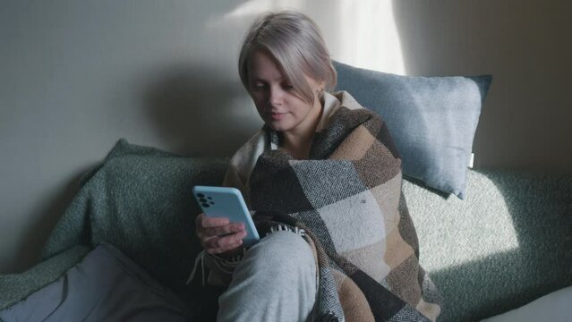 Girl under cozy blanket swiping typing message on smartphone sitting on couch.
