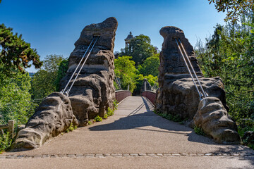Paris, France - 01 15 2023: Park des Buttes Chaumont. View of the Footbridge joining the belvedere island, the Temple of the Sibyl and buildings behind. - obrazy, fototapety, plakaty