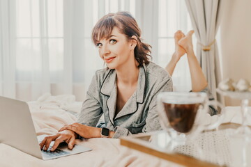 Freelancer woman wears pajamas, she lies in bed and uses work on laptop, computer, study on the...