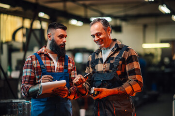 Two factory workers reading measures on a caliper and writing on a clipboard.