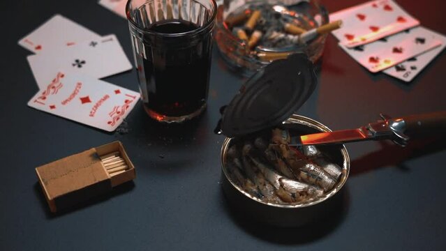 Still life with a glass of wine, a smoking cigarette, playing cards and an open can of fish. Playing cards at the table, against the background of food, wine and cigarettes.