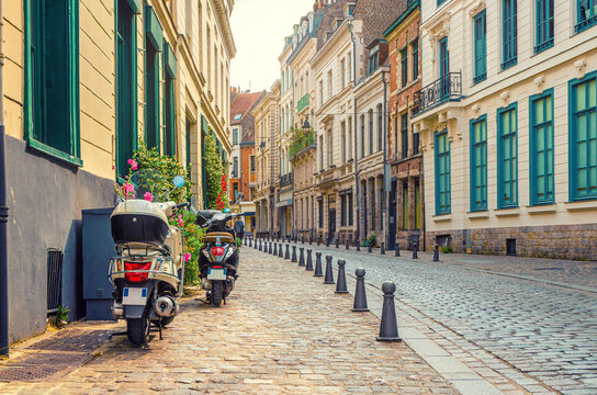 Two scooters bikes parked on empty narrow cobblestone street, paving stone road, old colorful buildings with stone walls in Lille old town historical city centre, French Flanders, Northern France