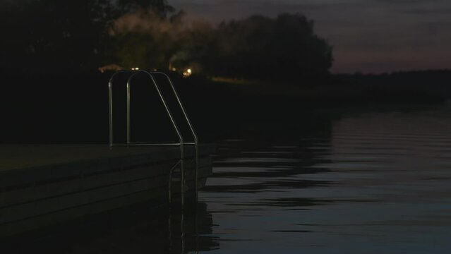 A night shot with a ladder that descends from the pier into the water. Swimming at night in the river. Smoke from the bathhouse is visible in the background. High quality 4k footage