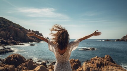 Woman with outstretched arms enjoying the wind and breathing fresh air on the rocky beach, back shot, summer time - obrazy, fototapety, plakaty
