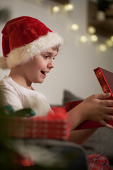 Obraz na płótnie Canvas Joyful boy in a Santa hat opens a Christmas present. Boxing day. Christmas Eve Magic night