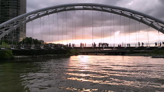 Arch bridge at Humber Bay, cloudy sunset, people walking and riding bicycles, on the pedestrian bridge with sunset peaking from behind them. Drone footage