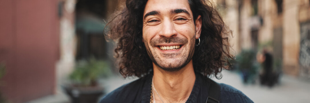 Young attractive italian guy with long curly hair and stubble looks into the camera and smiles at old buildings background. Stylish man with an earring in his ear and lot of chains