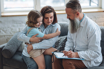 Doctor check heart during regular checkup in hospital. Sick little girl receive medical care. Senior male paediatrician examining sick kid sitting on mother's lap with stethoscope during home visit. 