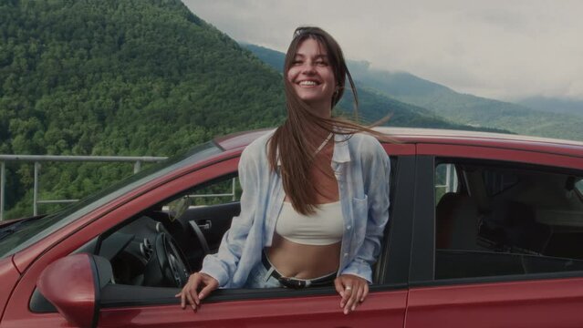 Portrait: Beautiful girl in a red car leans out through an open window, smiling. Pretty woman enjoys traveling and roadtrip. Summer wonderlust in the mountains. Active lifestyle in nature.