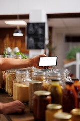 Client in zero waste shop using mockup mobile phone to analyze pantry staples. Vegan customer thoroughly checking local supermarket food items are allergens free with isolated screen smartphone - obrazy, fototapety, plakaty