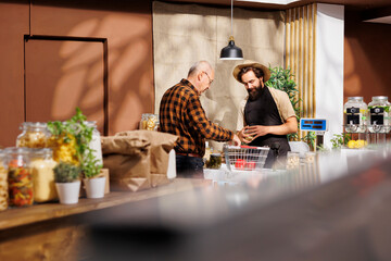 Elderly man at checkout in zero waste shop, buying locally grown ecofriendly vegetables from friendly storekeeper. Older customer purchasing organic chemicals free groceries