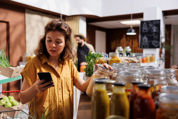 Woman in local groceries store using smartphone notes app to check shopping list. Meticulous customer looking to replenish pantry at home with organic eco friendly food essentials