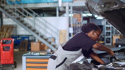African american woman in repair shop servicing broken vehicle using professional tools, requesting help from more experienced coworker. Mechanic in car service assisting colleague to fix automobile