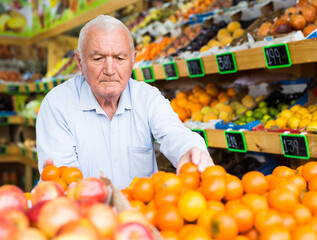 European old man choosing mandarins while standing in salesroom of greengrocer.