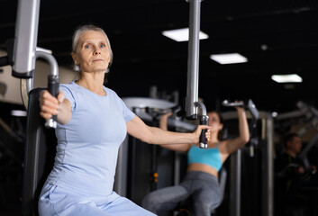 Fototapeta na wymiar Elderly female training trains muscles of arms and press on the simulator in gym