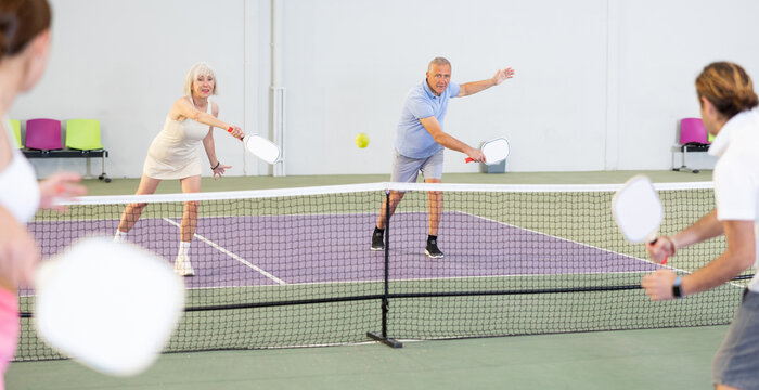 Sporty Couple Of Senior Woman And Man Playing Doubles Pickleball On Indoor Court, Ready To Hit Ball. Sport And Active Lifestyle Concept..