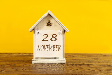 Calendar for November 28: the name November in English, numbers 28 on a decorative house on a wooden table, yellow background