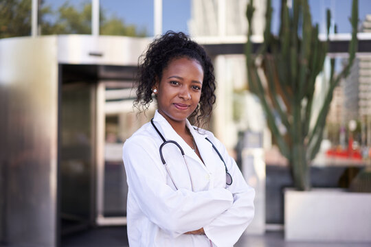 Looking at the camera portrait of a young confident woman doctor. Health professional outside the hospital. Crossed arms African American cheerful female health care worker. Copy space