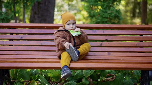 Baby boy wearing warm clothes sits on the bench outdoors. Kid holds himself by the shoe and drinks fruit puree from doypack.