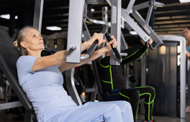 Fototapeta na wymiar Concentrated aged woman leading healthy active lifestyle doing strength training in gym, performing chest press in exercise machine