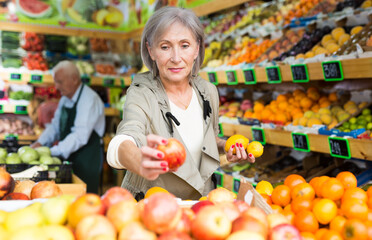 Mature woman standing amongst sheves with fruits and vegetables and selecting fruits. Senior man worker setting out goods in background.