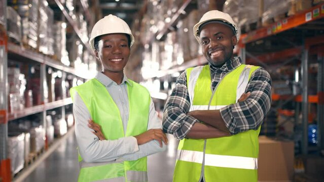African American woman and man in protective uniform looking at each other at warehouse. Two industrial workers smiling at background of multi tiered shelves. Woman and man with arms crossed.