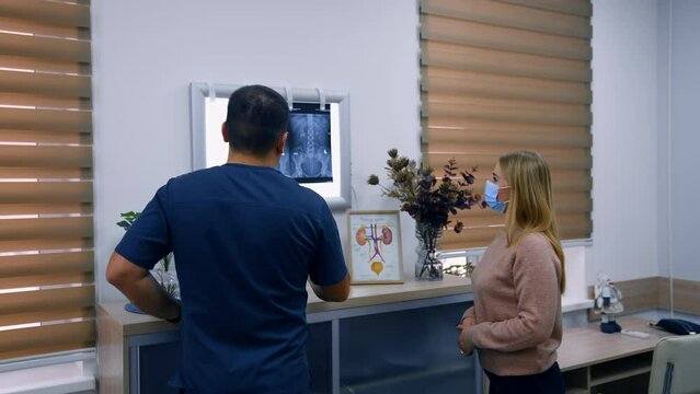 Patient and doctor standing in the hospital cabinet. Medic explaining the x-ray scan hanging on the light screen.