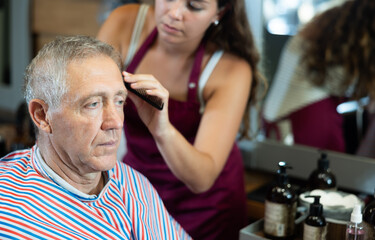 Relaxed elderly man wearing salon cape sitting in barbershop, getting haircutting by professional female barber using clippers..