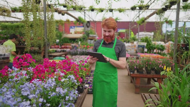 Young Man in Green Apron Utilizing Tablet for Inventory at Flower Shop. Male Employee Engaging with Modern Tech in Retail Environment