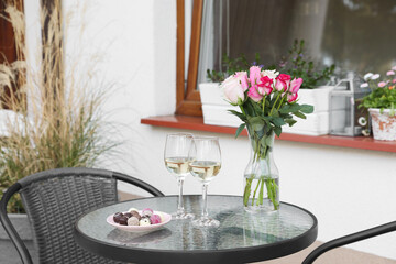 Vase with roses, glasses of wine and candies on glass table near house on outdoor terrace