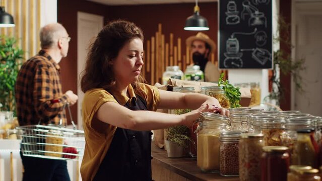 Portrait of smiling zero waste supermarket owner using biodegradable glass containers for its bulk food to lower climate impact. Cheerful woman in local grocery shop arranging merchandise