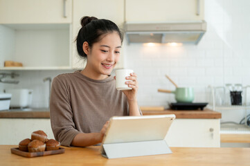 A woman is working from home, sipping her coffee and using her tablet at a table in the kitchen.