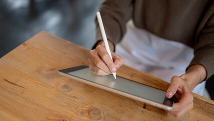 A woman holding a stylus pen and drawing or writing on a digital tablet at a table in a coffee shop.