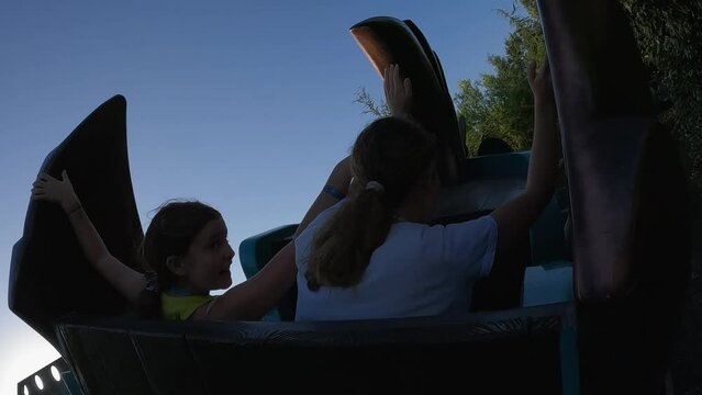 Young girls with arms raised have fun riding adrenaline twisting roller coaster at amusement park. Slow motion