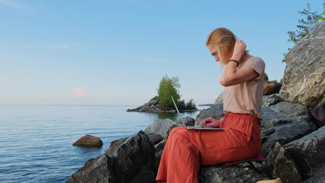A digital nomad, blonde caucasian woman sits by the sea and works remotely. Female freelancer uses laptop computer on empty seashore. Office for introvert outdoor.