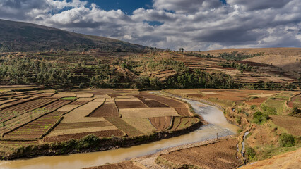 The river winds through the valley. Cultivated agricultural fields are located on the banks. Rice terraces rise up the hillside. Mountain on the background of blue sky, clouds. Madagascar.