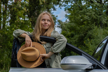 Blonde woman in hat staying next to car door. Young tourist explore local travel making candid real...