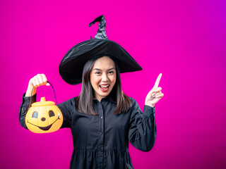 Portrait of an Asian Indonesian woman wearing a Halloween-themed costume with a witch hat, pointing towards something with her hand while holding a pumpkin. Isolated against a magenta background.