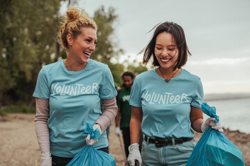 Two female volunteers in uniforms talking and carrying waste by the river