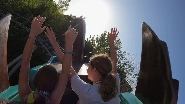 Two young girls with arms raised have fun riding adrenaline twisting roller coaster at amusement park. Slow motion