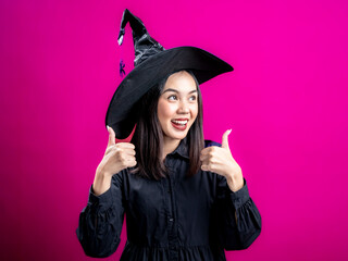 Portrait of an Asian Indonesian woman wearing a Halloween-themed costume with a witch hat, expressing agreement with a thumbs-up gesture. Isolated against a magenta background.