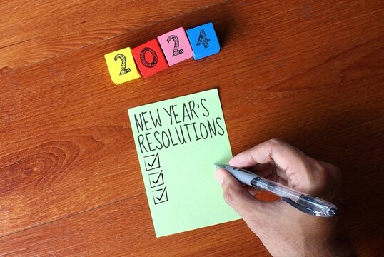 Top view image of wooden cubes with number 2024 and a person writing New Year's resolutions on a sticky note.