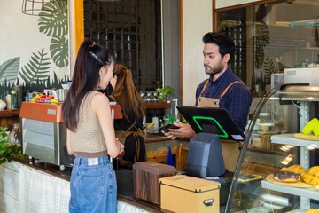 Young Indian barista or cashier worker man hold payment machine accept credit card cashless transaction from Asian customer woman, modern technology paying consumer eatery product cafe and bakery shop