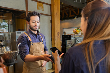 Young Indian baristas or cashier workers man holding a payment machine for scan, cashless technology and credit card payment concept. Customer using smartphone for payment to owner at cafe restaurant