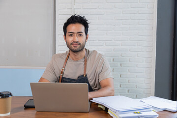Handsome Indian male restaurant owner sit and looking at camera with confidence. Man employee wearing apron calculate account income and expenses of shop, personal business concept.