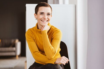 Young Modern Woman with Short Hair, Sitting in an Office Chair, Smiles Radiantly with Chin Resting