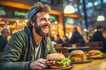Muurstickers Cheerful hipster man eating tasty burger and having fun while gathering with friends in open air bar at evening. Fast food eating on outside food court. Concept of pleasant past time with friends © KRISTINA KUPTSEVICH