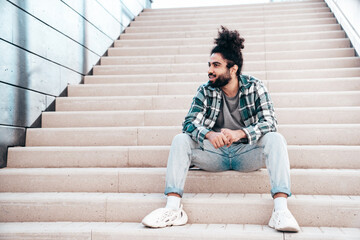 Handsome smiling hipster model. Unshaven Arabian man dressed in summer casual clothes, jeans and shirt. Fashion male with long curly hairstyle posing in the street. Sitting at the stairs